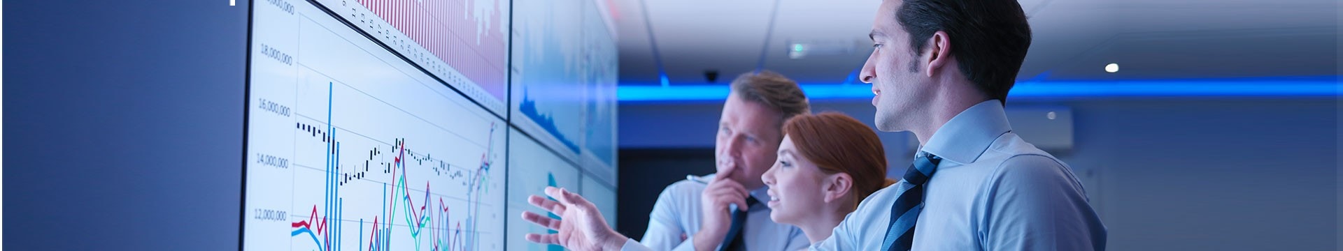 Three business people discuss graphs on screen in meeting room  