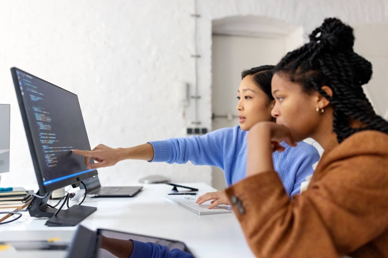 Two women pointing at code on computer monitor